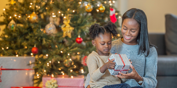 A little girl sitting on her mom's lap at Christmas opening a gift.