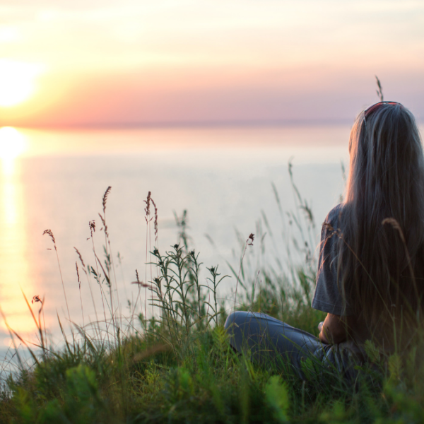 A woman sitting on the banks for a pond looking at the sunset.