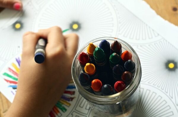 a preschooler's hand coloring a coloring sheet, with a jar of crayons sitting nearby - while learning at home.